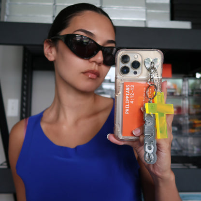 Woman holding a phone case with a Kainos keychain, cross keychain, and scripture, wearing sunglasses.